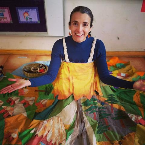 Una mujer sonriente viste un vestido colorido que se extiende sobre el suelo, rodeada de naturaleza.