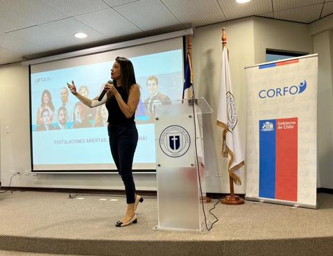 Una mujer presenta un discurso en un evento mientras se proyectan imágenes en una pantalla detrás de ella.