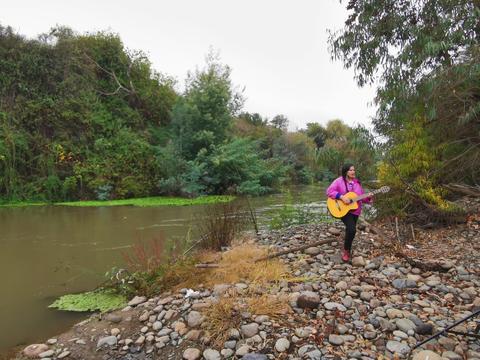 Una mujer toca la guitarra al lado de un río rodeado de vegetación.