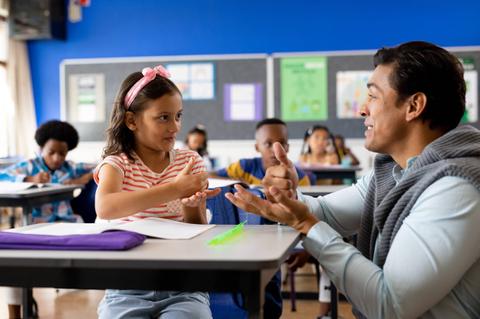 Un profe y una niñita interactúan en clase mientras otros compañeros los observan.