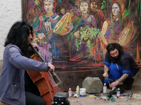 Dos artistas trabajan en un espacio creativo, uno toca el violonchelo mientras el otro pinta frente a un mural colorido.