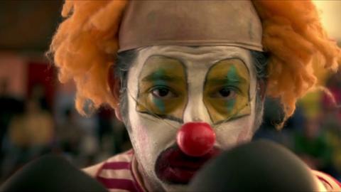 Un payaso con rostro pintado y peluca colorida mira a cámara.