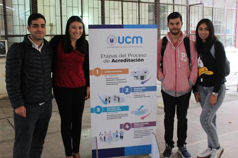 Un grupo de cuatro jóvenes se encuentra frente a un cartel sobre las etapas del proceso de acreditación de la UCM.