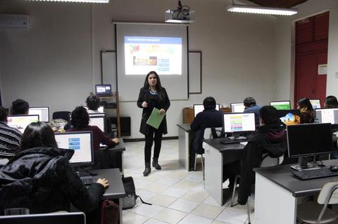Una mujer está dando una clase en un aula con computadoras mientras los estudiantes la escuchan atentamente.