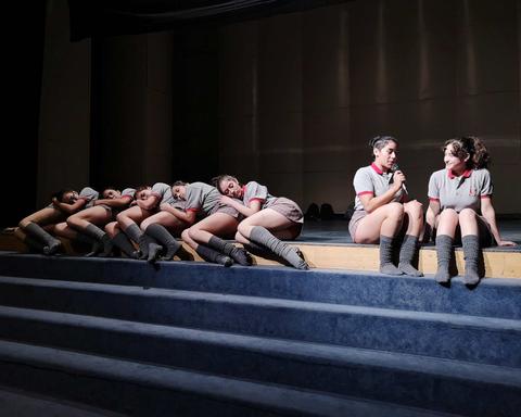 Un grupo de bailarinas en un escenario, algunas sentadas y otras recostadas, mientras dos de ellas conversan.