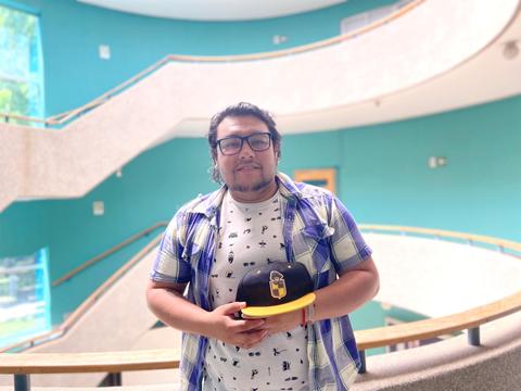 Una persona sonriente sostiene una gorra en un entorno arquitectónico moderno.