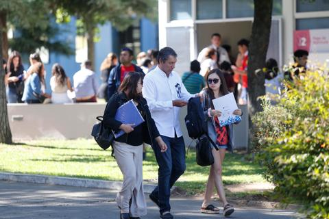 En la imagen, tres personas caminan por un campus universitario rodeadas de estudiantes.