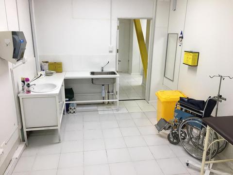 La imagen muestra un cuarto de hospital con un lavabo, una silla de ruedas y un área de tratamiento vacía.