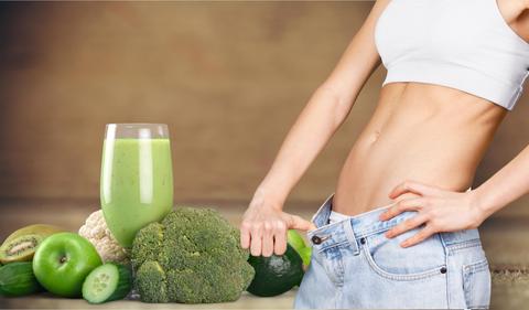 Una mujer muestra su cintura delgada junto a un batido verde y verduras saludables.