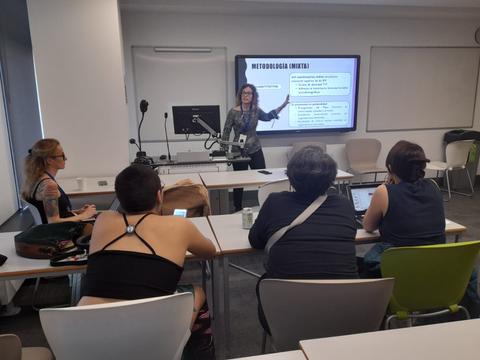 Una mujer está presentando en una sala de clases mientras un grupo de personas la observa.