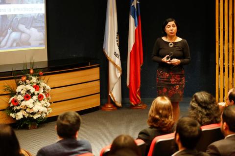 Una presentadora habla frente a una audiencia en un evento con decoraciones y una pantalla proyectora.