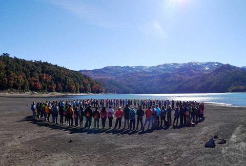 Un grupo numeroso de personas se reúne en forma de círculo en una playa junto a un lago rodeado de montañas y árboles con follaje de otoño.