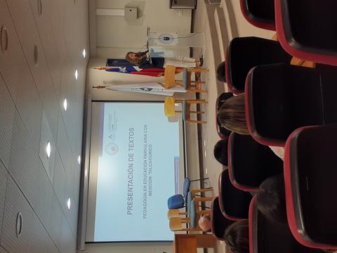 Una mujer presenta un texto en un auditorio con banderas al fondo.