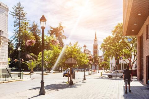 La imagen muestra una calle arbolada con farolas y edificios, iluminada por la luz del sol.