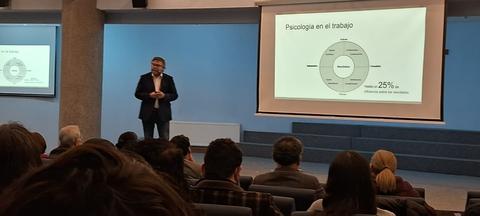 Un expositor presenta sobre psicología en el trabajo en una sala llena de personas.