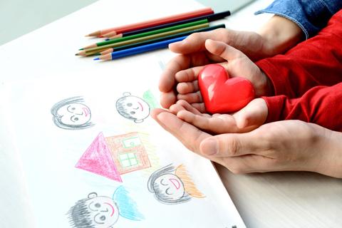 Manos sosteniendo un corazón rojo sobre un dibujo infantil de caras y una casa.