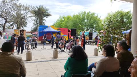 Una orquesta se presenta al aire libre, mientras un público curioso observa desde la distancia.