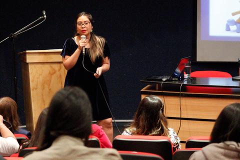 Una mujer presenta en un escenario mientras los asistentes la observan desde asientos.