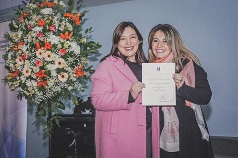 Dos mujeres sonrientes sostienen un certificado frente a un arreglo floral.