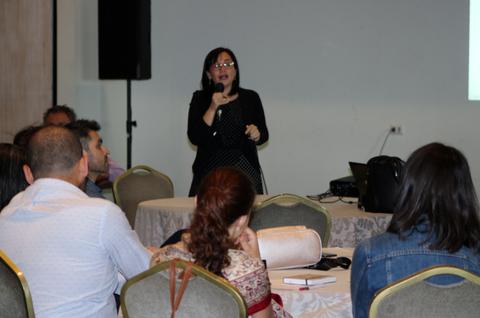 Una mujer habla en público frente a un grupo de personas sentadas en un ambiente de conferencia.