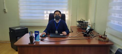 Un hombre sentado en un escritorio con una computadora, usando mascarilla, en una oficina bien iluminada.