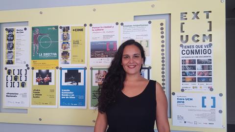 La imagen muestra a una mujer sonriendo frente a un mural con carteles de eventos culturales y cinematográficos.