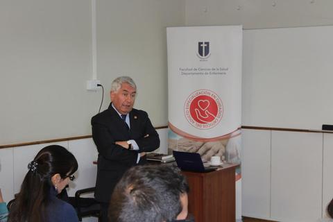 Un hombre mayor está dando una charla en un salón con un banner de la facultad de Ciencias de la Salud.