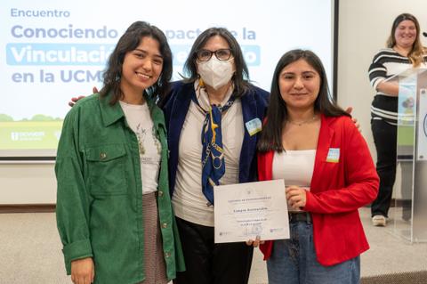 Tres mujeres posan juntas en un evento, sosteniendo un certificado, mientras una cuarta persona aparece detrás sonriendo.