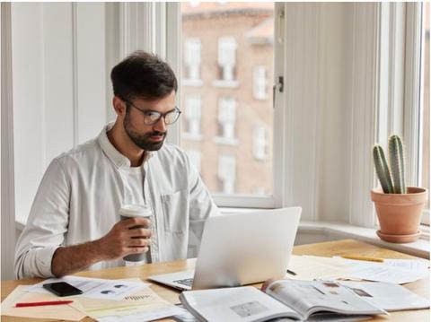 Un hombre trabaja frente a una computadora portátil mientras sostiene una taza de café en un entorno con luz natural.