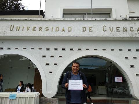 Una persona sostiene un certificado frente a la Universidad de Cuenca.