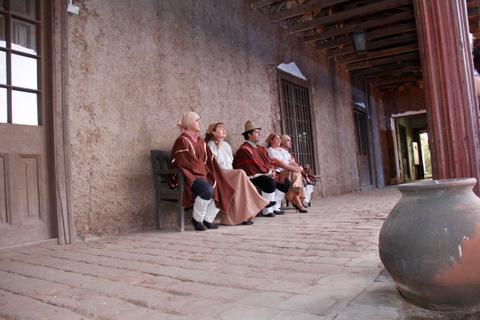 Un grupo de personas vestidas con trajes tradicionales se sienta en un banco frente a una pared de adobe.