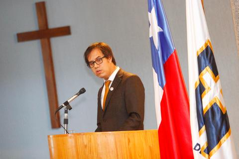 Un hombre habla en un podio frente a banderas chilena y de una institución, con una cruz en la pared detrás.