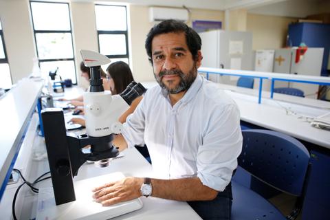Un hombre con barba sonríe mientras se sienta frente a un microscopio en un laboratorio.