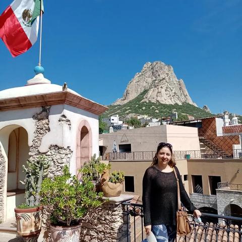 Una mujer sonriente posa frente a un paisaje montañoso con una bandera de México ondeando al fondo.