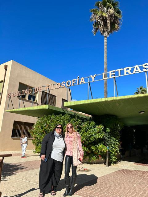 Dos mujeres posan frente al edificio de la Facultad de Filosofía y Letras bajo un cielo azul claro.