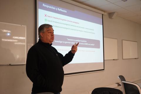 Un hombre está presentando en una sala de clases mientras apunta hacia una diapositiva en la pantalla.