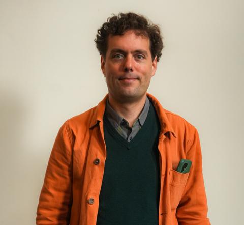Un hombre con una chaqueta naranja sonríe frente a un fondo claro.