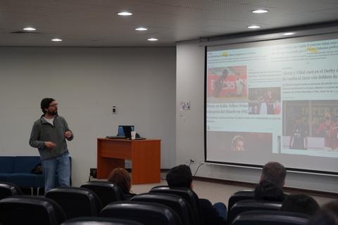 Un tipo está dando una charla en una sala con una pantalla grande que muestra varias imágenes y textos.