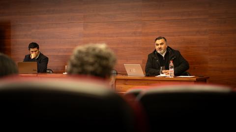 Una reunión en un salón con dos hombres, uno hablando y otro escuchando desde una computadora.