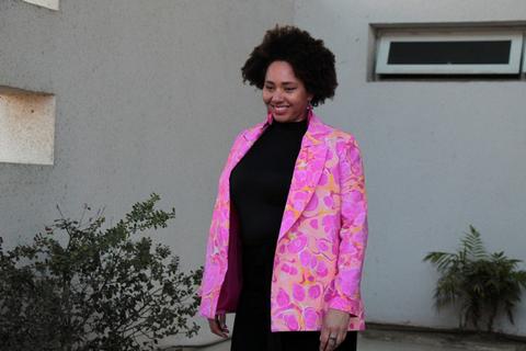 Una mujer sonriente viste una chaqueta rosa con un diseño colorido y se encuentra de pie en un entorno exterior.