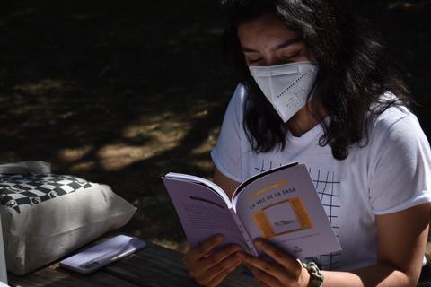 Una persona lee un libro sentada en una mesa al aire libre mientras lleva puesta una mascarilla.