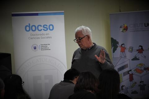 Un hombre mayor habla frente a un panel informativo en un evento relacionado con la ciencia social.