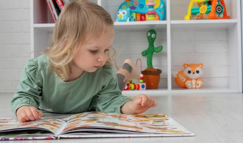 Niña pequeña leyendo un libro en el suelo rodeada de juguetes.