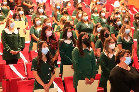 Un grupo de personas en un auditorio, la mayoría vestidas con uniformes verdes y utilizando mascarillas, se encuentran de pie mientras escuchan un discurso.