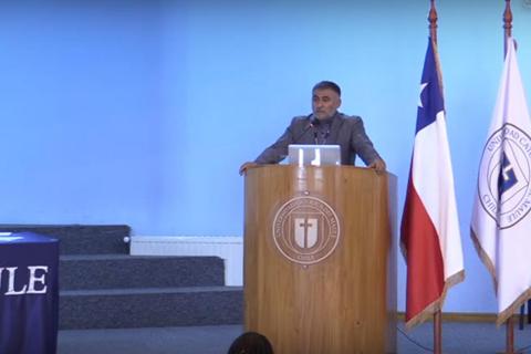 Un conferencista presenta en un escenario con banderas chilenas y un podio.