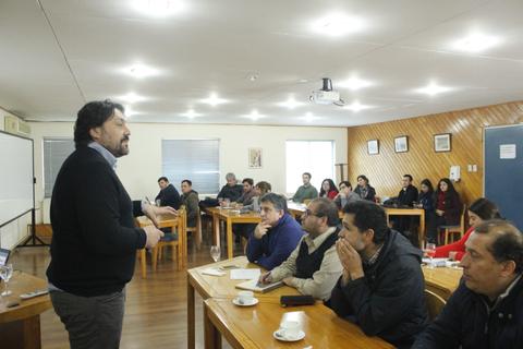 Una reunión en un salón con un ponente frente a un grupo de personas que lo escucha atentamente.