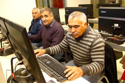 Tres hombres mayores están sentados en escritorios frente a computadores en un aula.