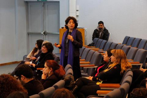 Una mujer con una bufanda azul presenta frente a una audiencia en un salón.