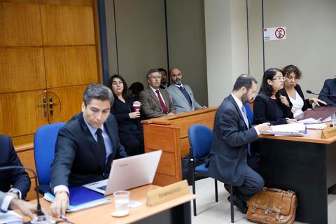 Un grupo de personas se encuentra en una sala de juicio, algunas sentadas y otras de pie, con documentos y computadoras en un entorno formal.