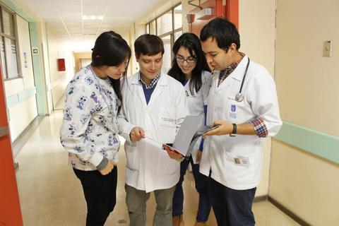 Cuatro jóvenes, dos reconocidos como médicos, revisan un dispositivo móvil en un pasillo de hospital.